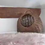 La vision de notre élevage, l'espace de mes chats | Coin douillet que j’ai aménagé pour mes chats avec un panier en osier placé contre un angle du mur, sous une poutre de bois rustique. Un tapis en fausse fourrure de couleur rose clair est étalé devant le panier sur l’un des plateaux transparents de la chatterie, ajoutant une touche de confort à l’espace.