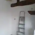 Les moments clés de l’aménagement de la chatterie | Présente une échelle grise en aluminium, portant des traces d’usure et de peinture, adossée à un mur blanc fraîchement peint. Des poutres en bois foncé sont apparentes, apportant une touche rustique à la pièce. Une prise électrique étanche munie d’un capot gris est visible sur le mur à côté de l’échelle. Le sol est recouvert de carreaux de couleur claire.