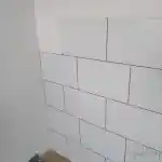 Les moments clés de l’aménagement de la chatterie | Cette image présente un mur partiellement recouvert de carreaux blancs. Les joints entre les carreaux sont bien visibles, indiquant que le travail de carrelage n’est pas encore achevé. La couleur blanche domine dans l’image, tant sur les carreaux que sur le mur. L’éclairage est naturel grâce à la grande ouverture de la chatterie, bien que la source lumineuse ne soit pas visible sur l’image.