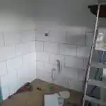 Les moments clés de l’aménagement de la chatterie | Cette photographie illustre la pièce dédiée à mes chats, en pleine rénovation. Des carreaux blancs sont partiellement installés sur le mur et le sol est revêtu de carreaux de couleur bois. La pièce est jonchée d’outils et de matériaux de construction. Un escabeau est également visible.